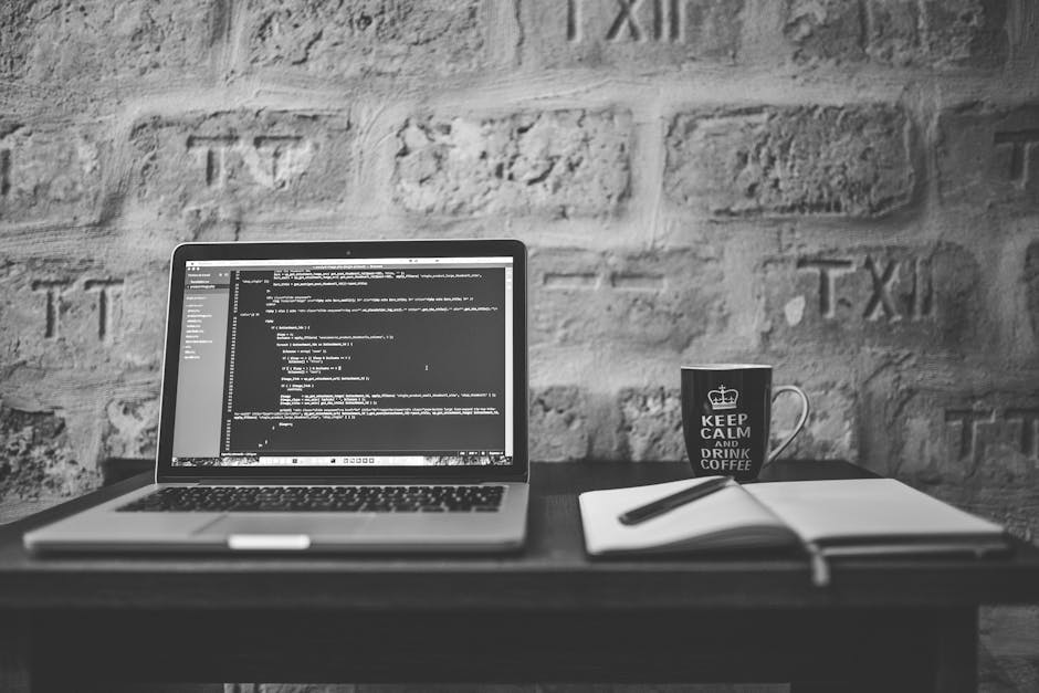 A black and white photograph of a workspace featuring a laptop open on a dark wooden desk displaying lines of code on its screen, a white notebook with a black pen resting on top, and a coffee mug wit