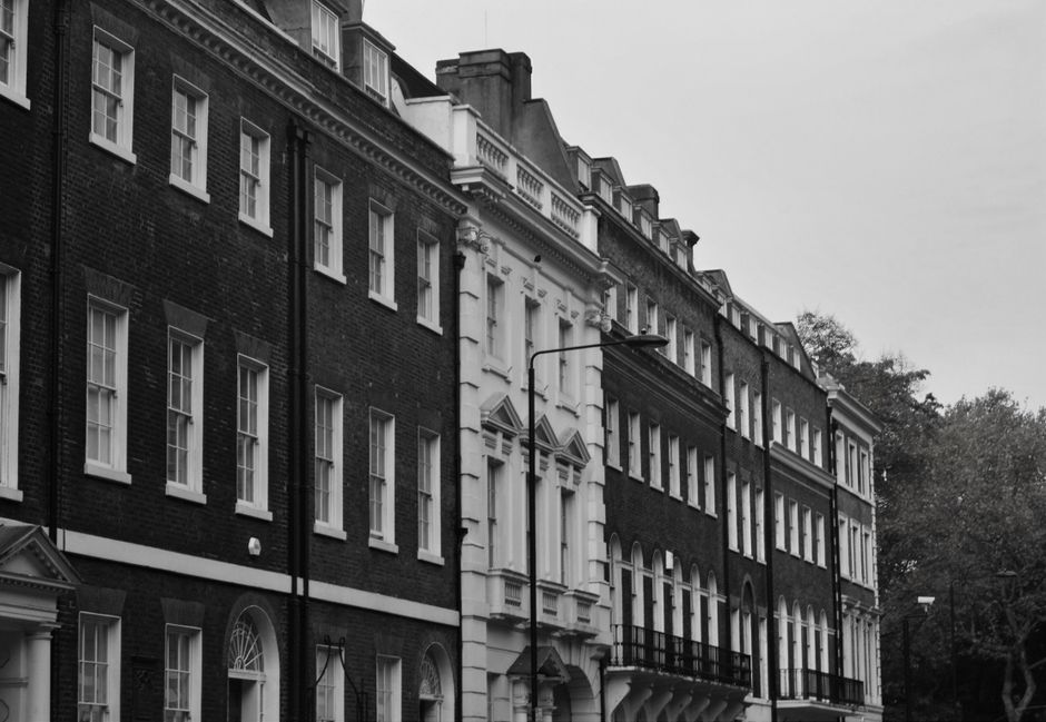 Photograph of a historic red-brick residential building situated on a city street, featuring large sash windows and small balconies with wrought iron railings. The building’s stone window surrounds