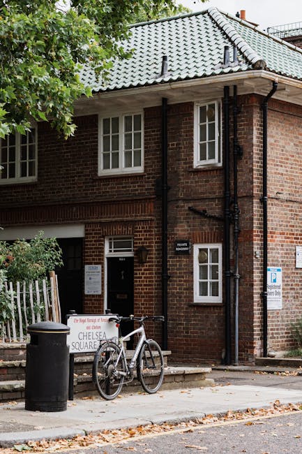Exterior view of a red brick residential building in SW3, featuring a tiled grey roof and white-framed double-glazed windows. The building's entrance includes a black door with a small sign, situated 